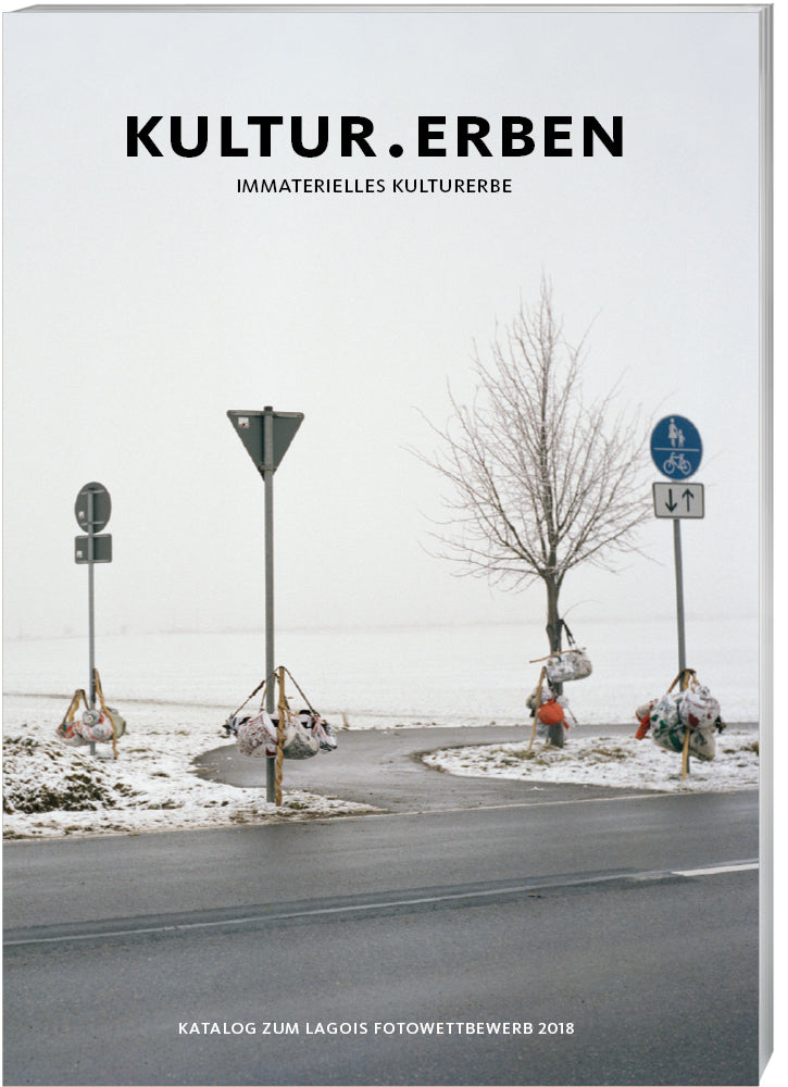 Cover des Katalogs "Kultur.Erben - Immaterielles Kulturerbe" mit einem winterlichen Landschaftsbild, das eine Kreuzung mit Verkehrsschildern und einem kahlen Baum zeigt.